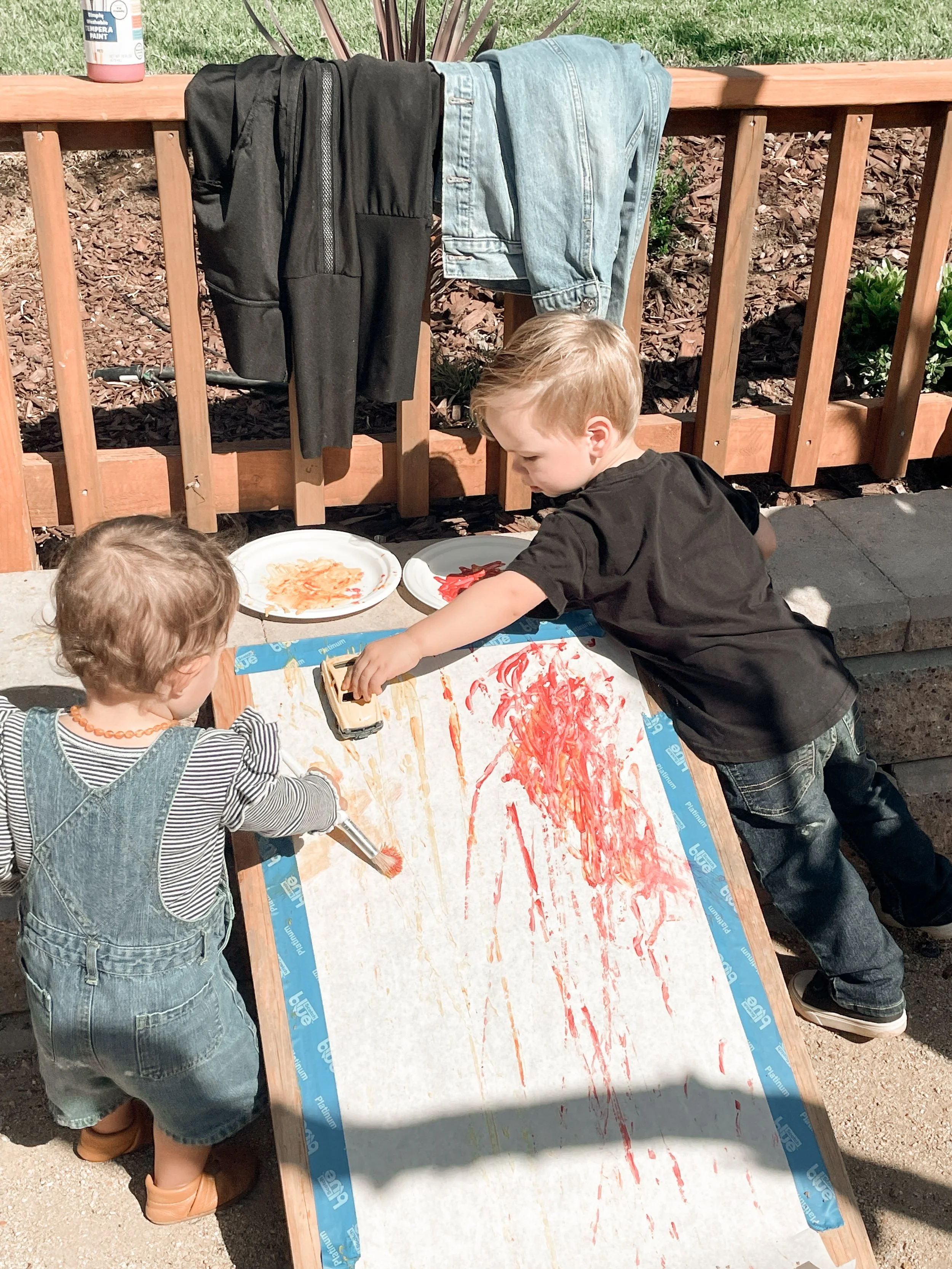 Three children playing outside, painting on a large sheet of paper attached to an easel. Two of the children are wearing denim overalls, and one is wearing a black t-shirt. One child is reaching for a toy car on the paper, which is covered in red and