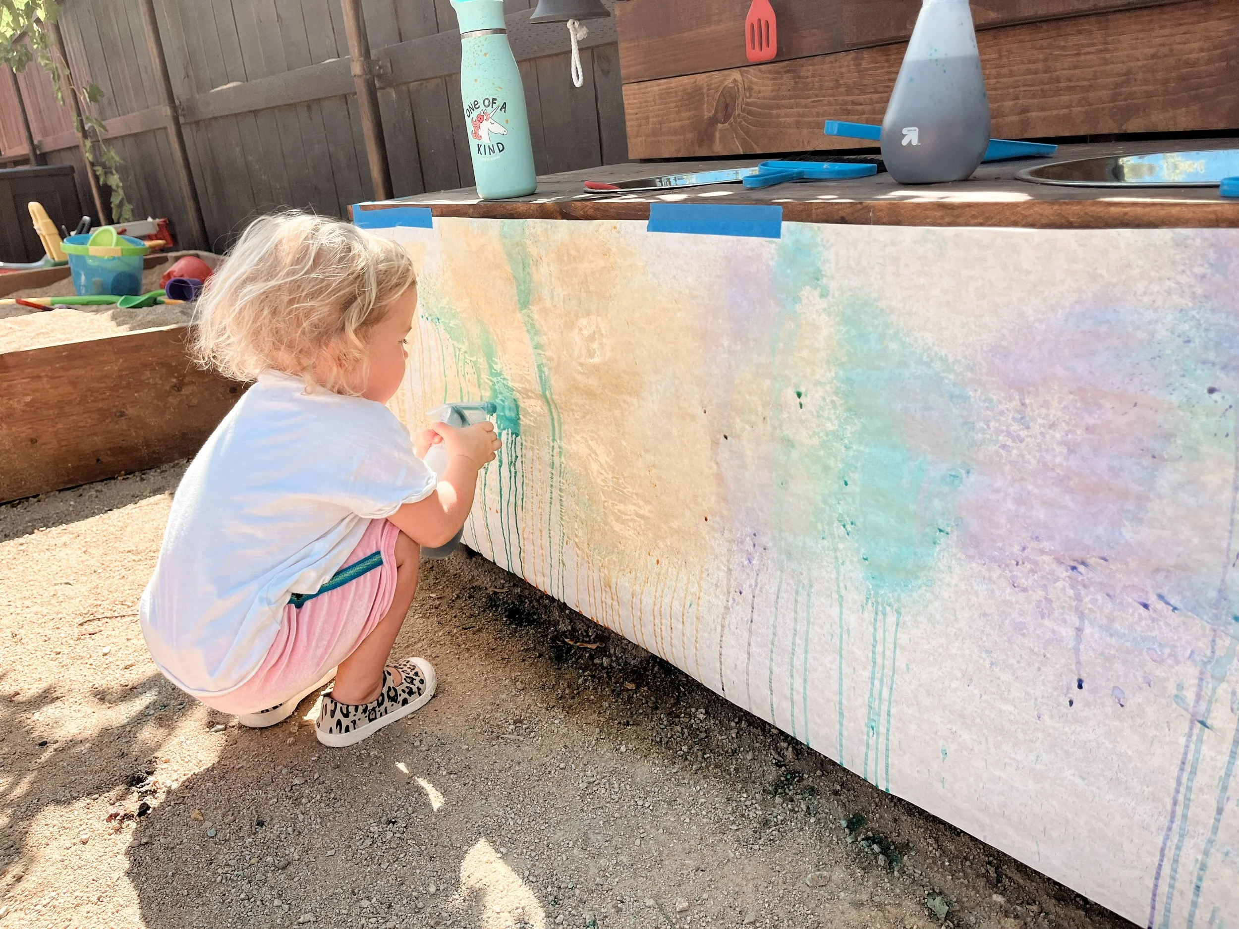 Young girl crouching down and painting a colorful mural on a large wooden surface outdoors, with various toys and gardening tools nearby.