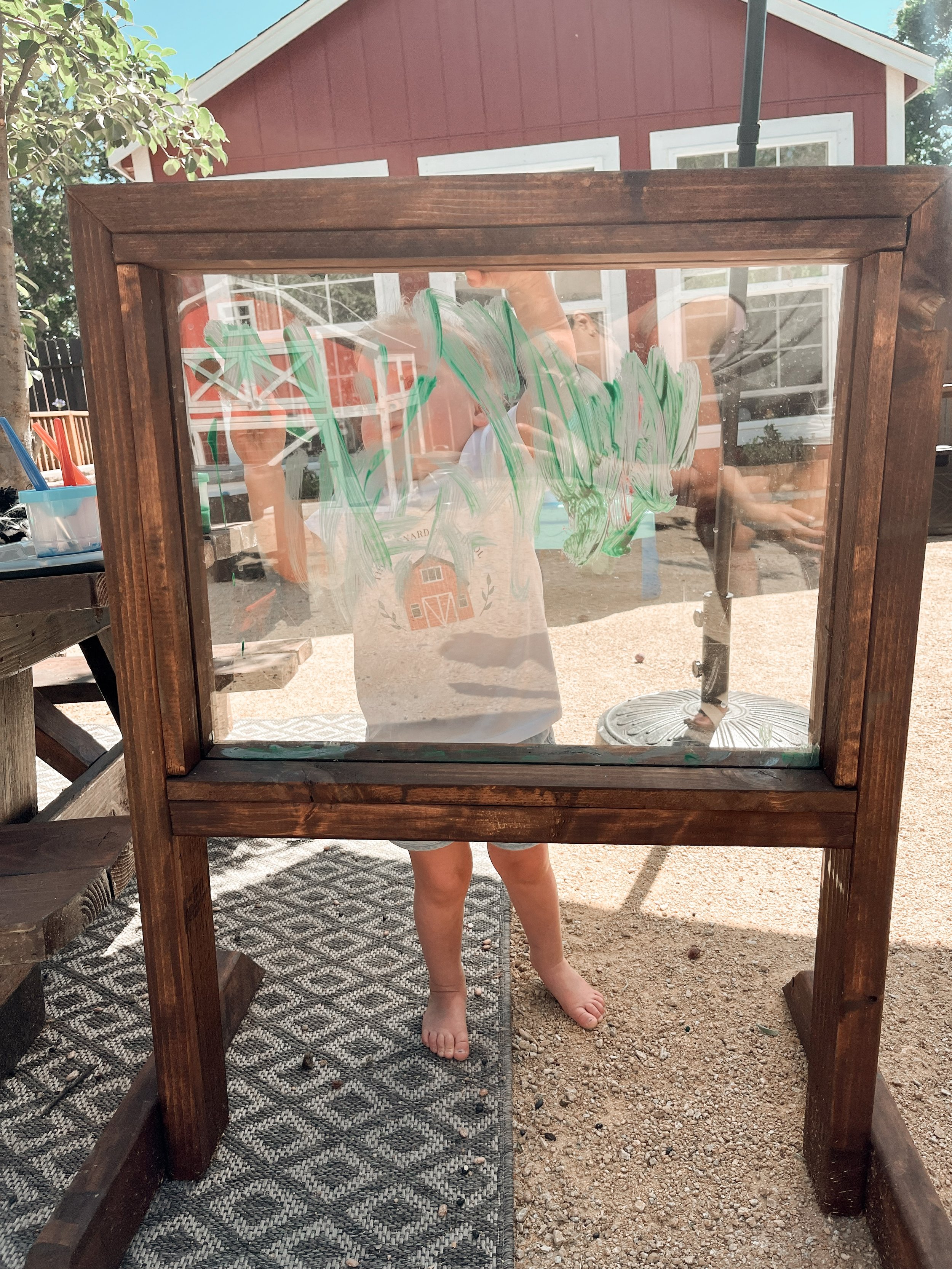 Child drawing on glass surface with green and white paint, view through wooden-framed glass, outdoor setting with house in background.