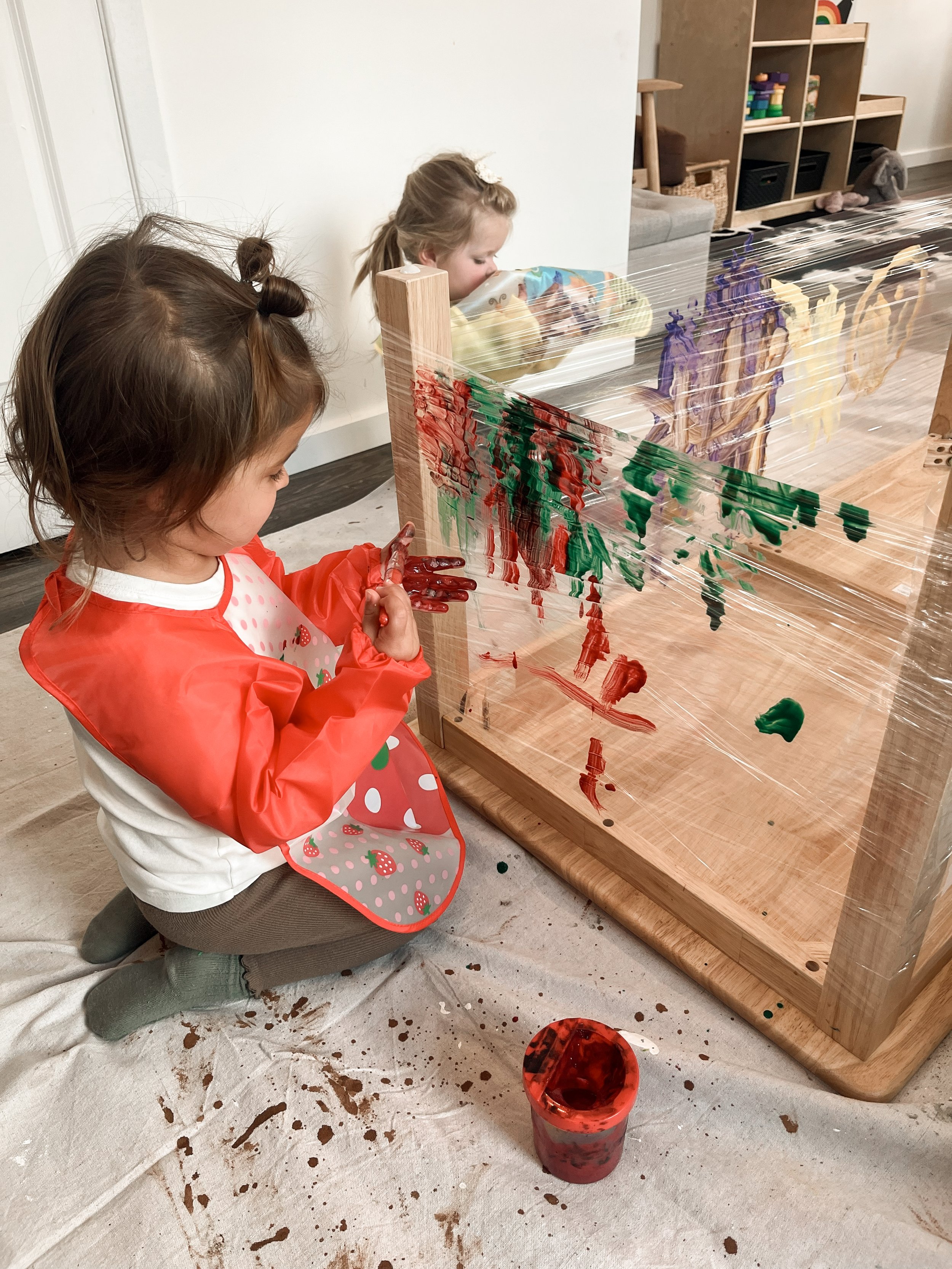 Two young girls are painting on a clear plastic sheet attached to a wooden frame in a room with a light-colored wall and shelves with toys and books in the background. The girl in the foreground is kneeling on a white cloth, wearing a red smock with 