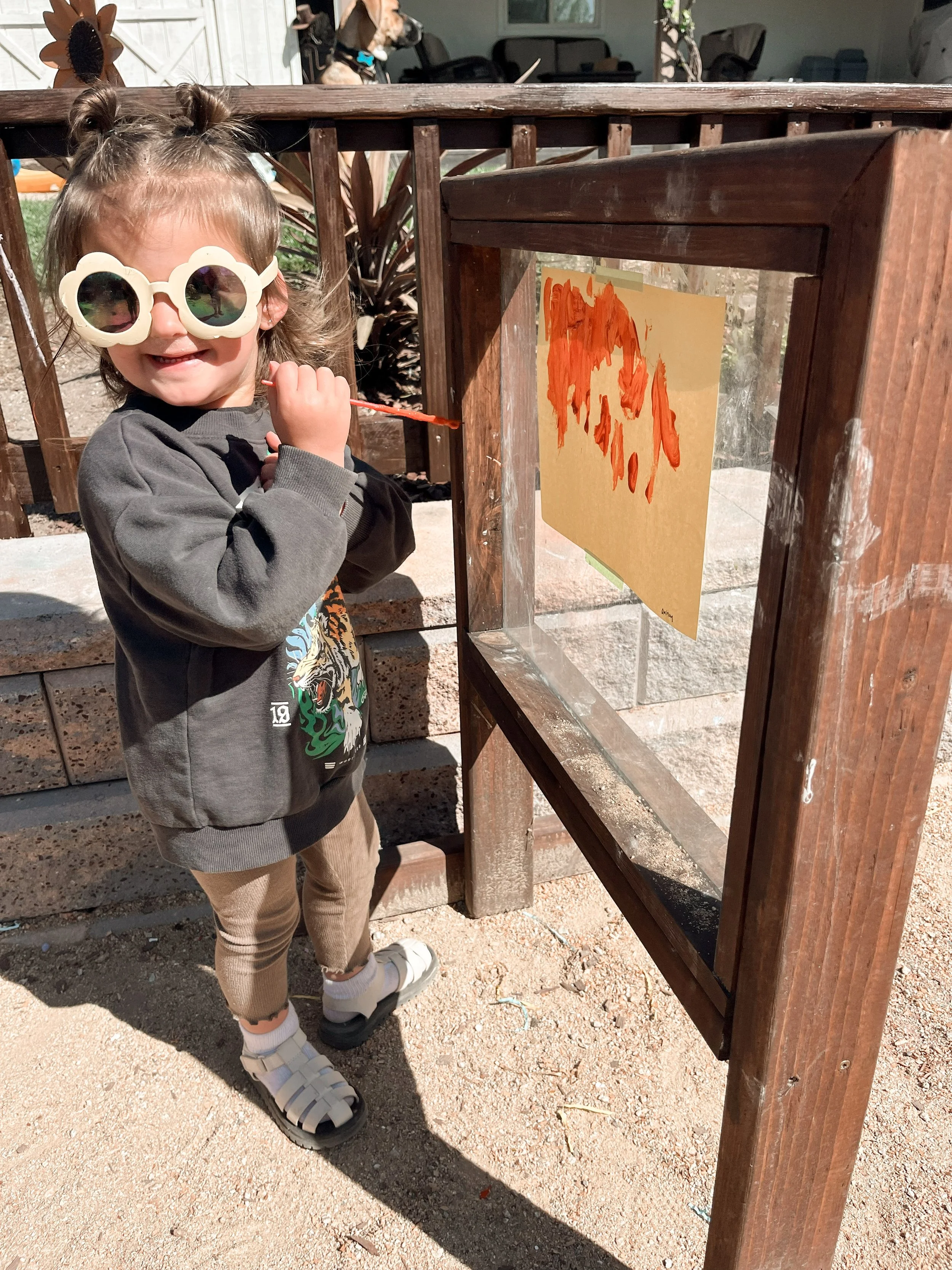 A young girl with short hair, wearing unique white sunglasses, a gray hoodie with a graphic print, tan pants, and white sandals, is standing outdoors near a wooden structure with an abstract orange painting taped to it. She is smiling and holding a s