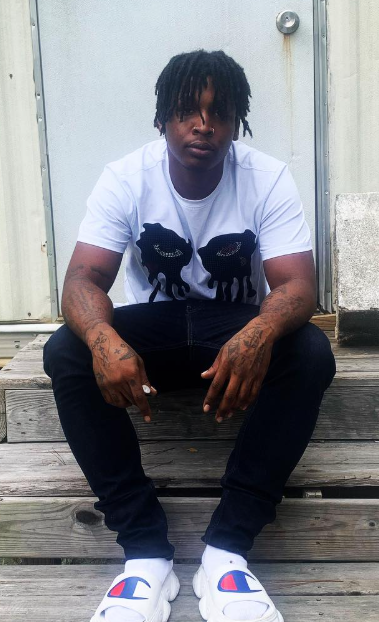 Young man sitting on wooden stairs, wearing a white t-shirt with black graphic, black pants, white sneakers, with tattoos on arms and dreadlocks.