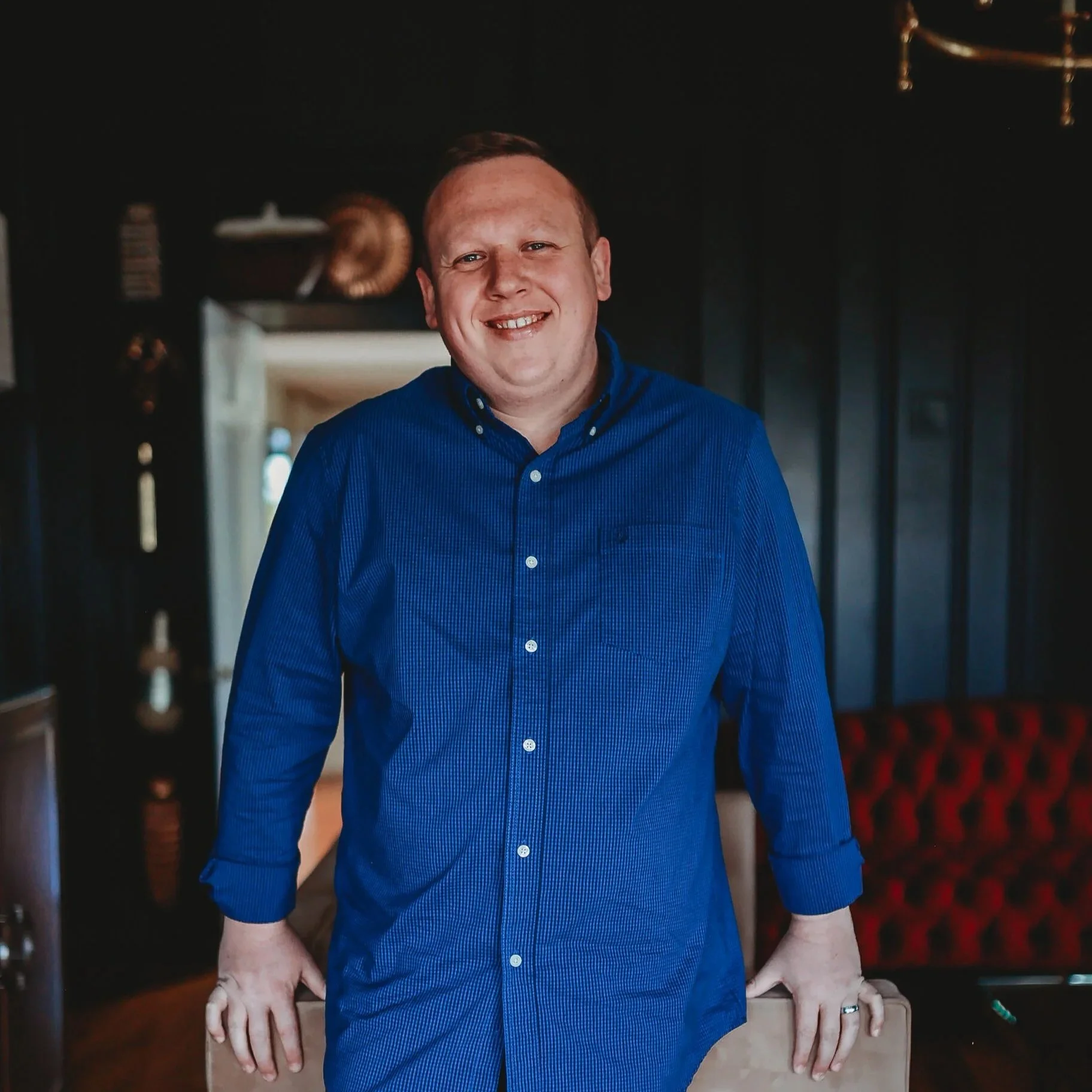 Smiling man in a blue shirt standing in a dimly lit room with dark walls and a red floral-patterned carpet, holding a cardboard box with his right hand.