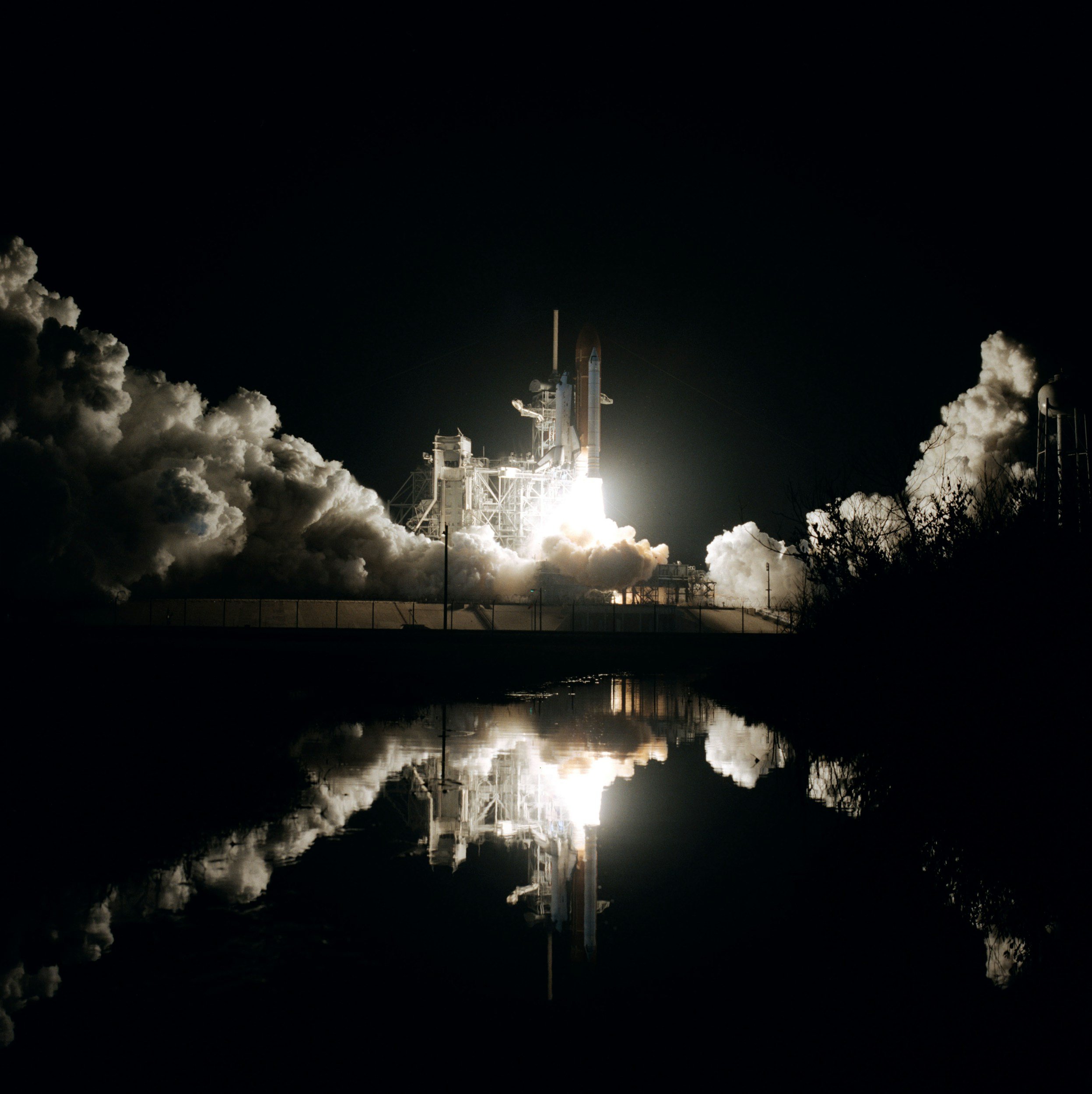 A rocket launching into the night sky surrounded by billowing smoke and clouds, with reflections on the water below.