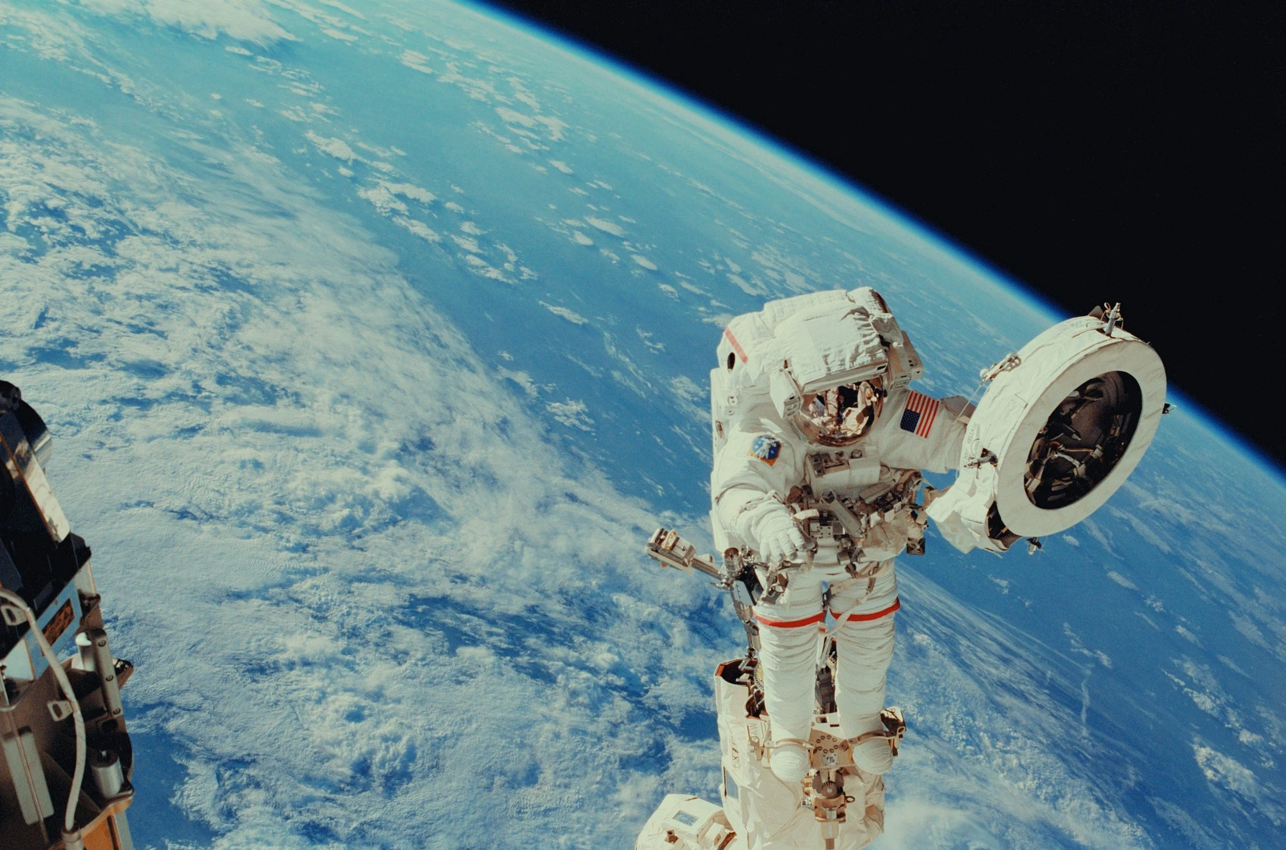 An astronaut in a white space suit performing a spacewalk outside a spacecraft with Earth visible in the background.