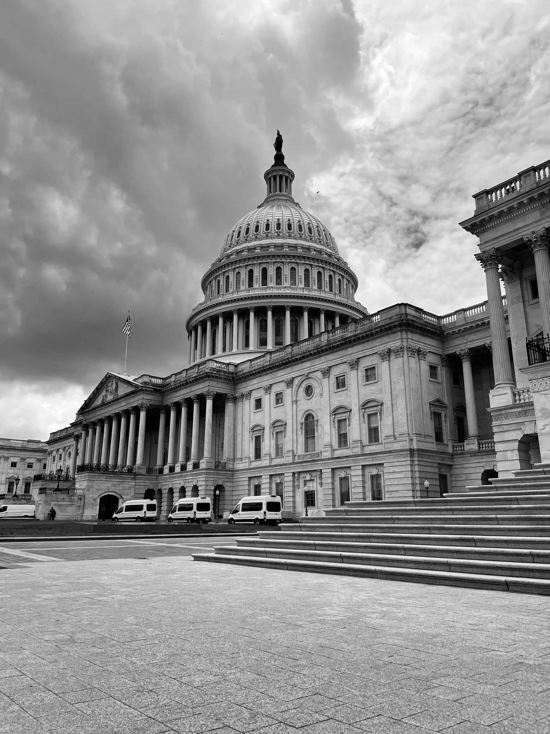 Black and white photo of the U.S. Capitol building with large steps, parked vans, and a flagpole. The sky is cloudy.