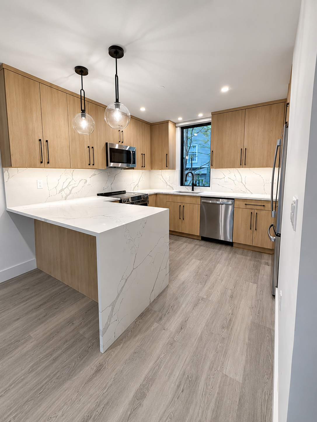 Modern kitchen with wooden cabinets, white marble countertops with gray veining, a stainless steel sink, microwave, oven, dishwasher, and a window above the sink.