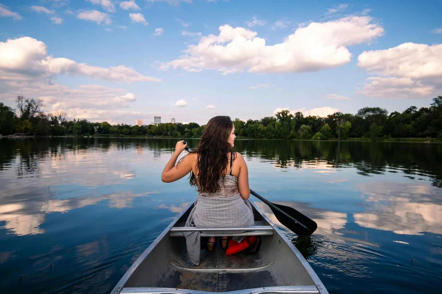 A woman with long brown hair paddling a canoe on a calm lake with greenery and trees on the shore and a city skyline in the distance under a partly cloudy sky.
