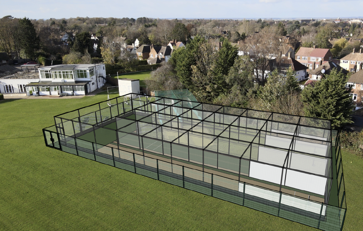 A large outdoor tennis court with glass walls and a black metal fence, situated on a grassy field with trees and residential houses in the background.