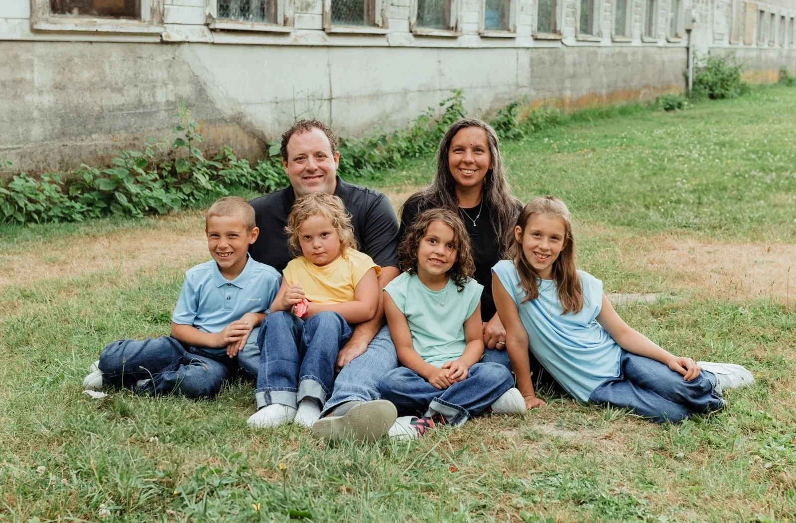 A family of seven posing outdoors on grass in front of a stone building with multiple windows. Adults are sitting behind four children who are seated or lying on the grass, smiling at the camera.