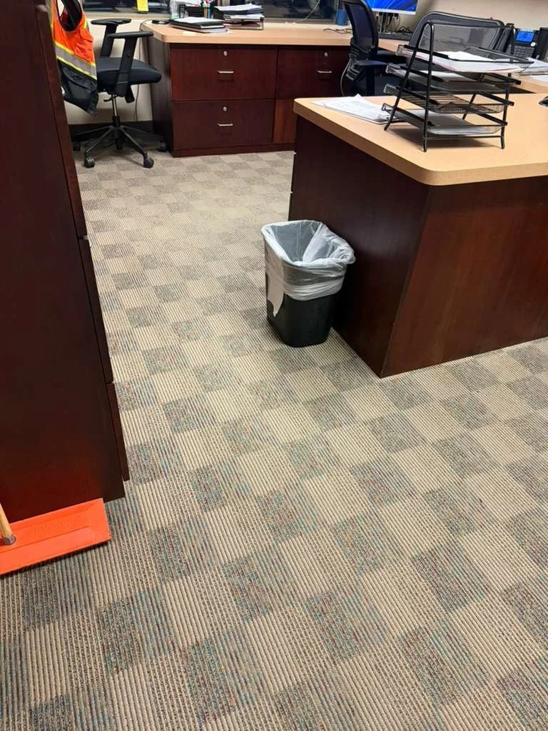 Espacio de oficina con escritorio, silla de oficina, bandejas para papeles, y una papelera.