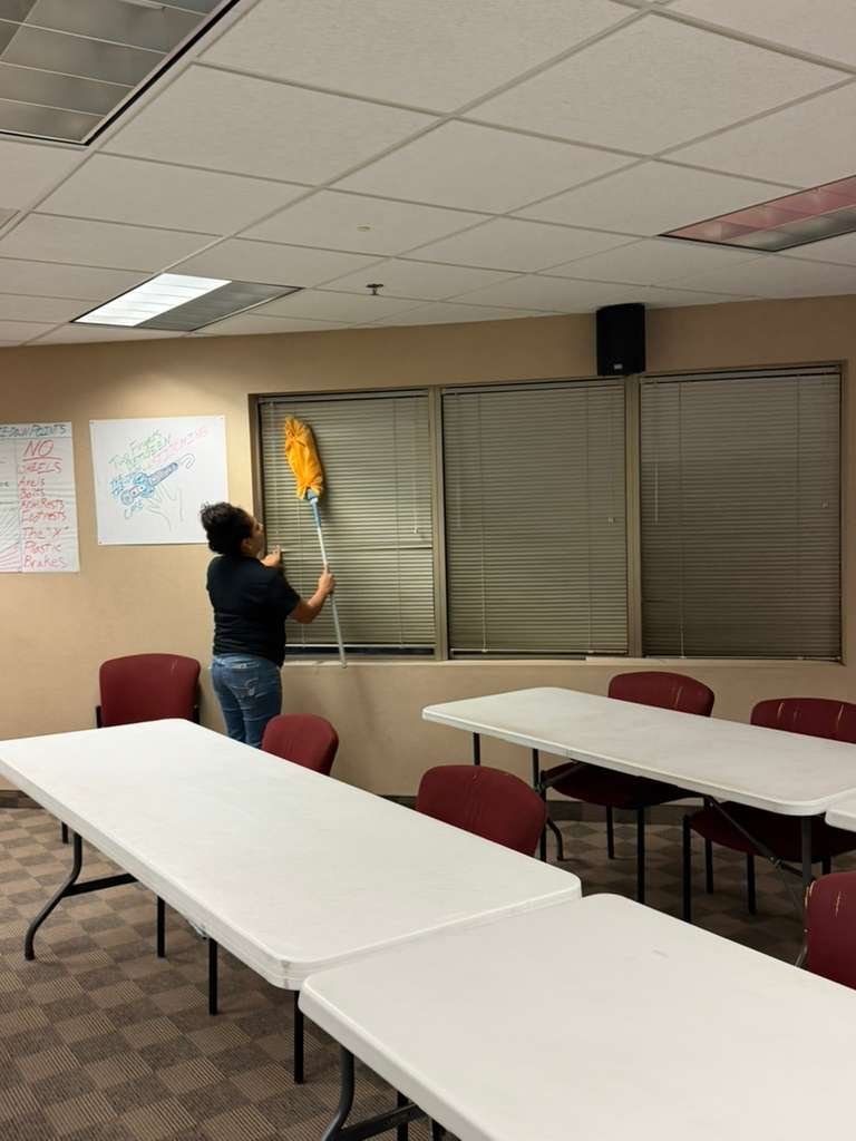 Persona limpiando ventanas con una escoba en una sala de reuniones con mesas y sillas rojas.