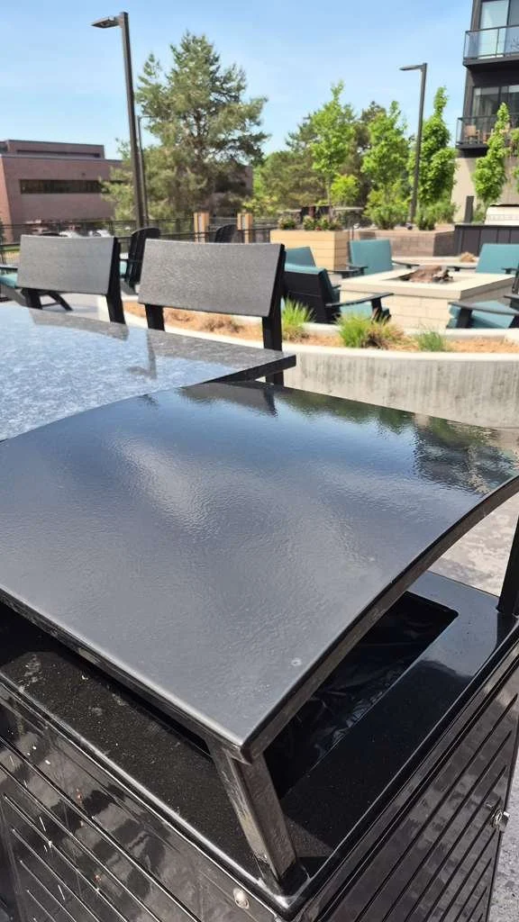 Mesa de exterior de acero con una tapa metálica negra y sillas alrededor, en un espacio al aire libre con áreas verdes y árboles.