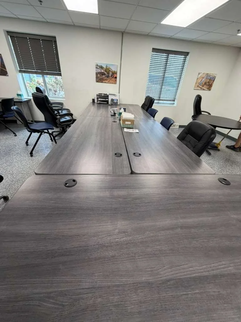Sala de reuniones con varias sillas y mesas de madera, ventanas con persianas, fotos en la pared y papelería en la mesa central.