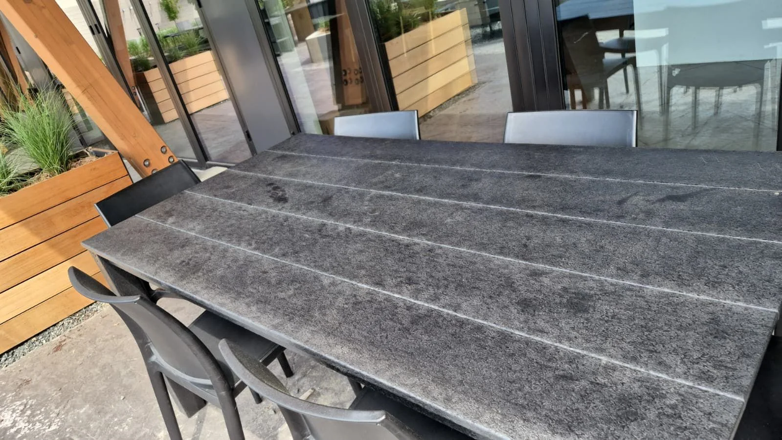 Mesa de piedra oscura en un espacio techado con sillas alrededor, ventanas con reflejos de vegetación, y paneles de madera.