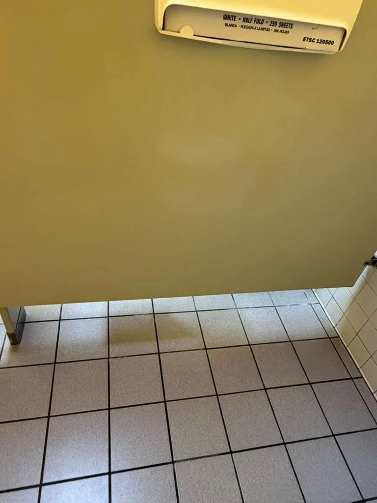 Una taquilla de papel higiénico en un baño público con pared beige y azulejos blancos y grises en el suelo.