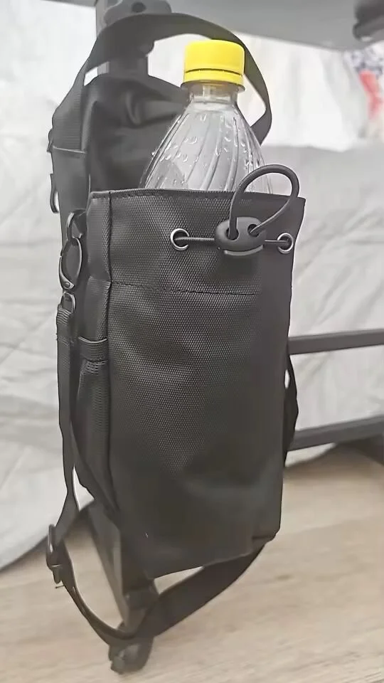 A black backpack with a water bottle that has a yellow cap in a side pocket, placed on a wooden surface near a bed or couch.
