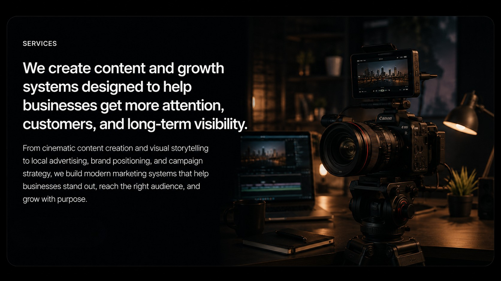 A professional camera setup on a desk, with a monitor and laptop displaying cityscape footage, in a dimly lit room with a lamp and small plants.