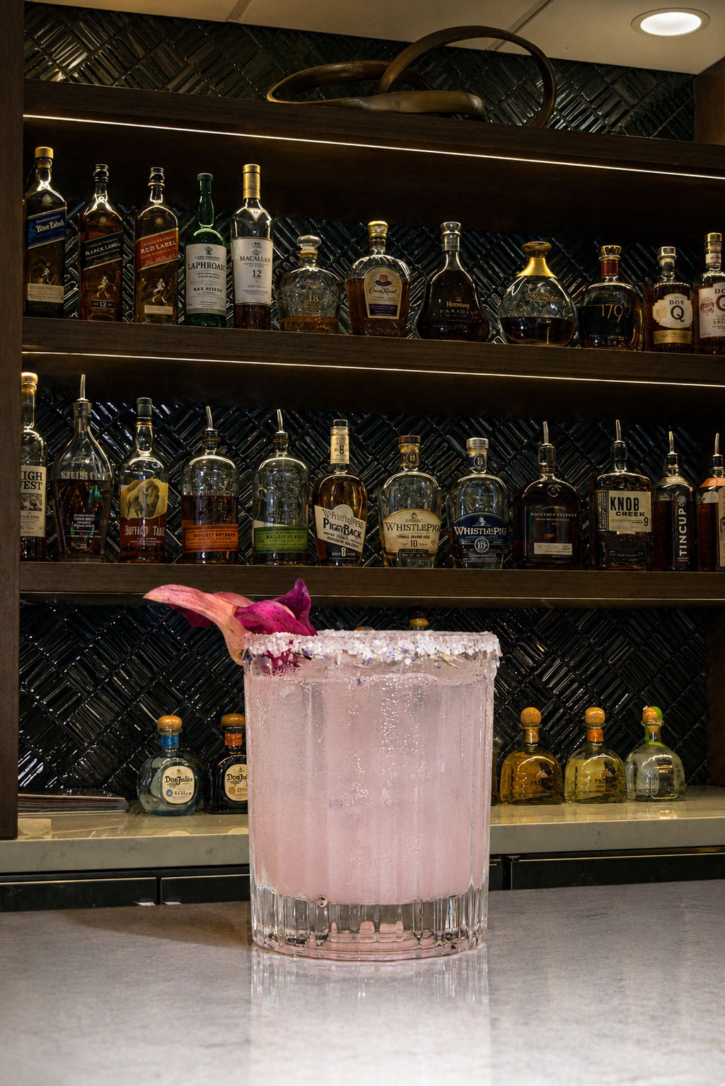 A pink cocktail garnished with a pink flower and a sugared rim, placed on a bar counter in front of a shelf filled with various bottles of liquor.