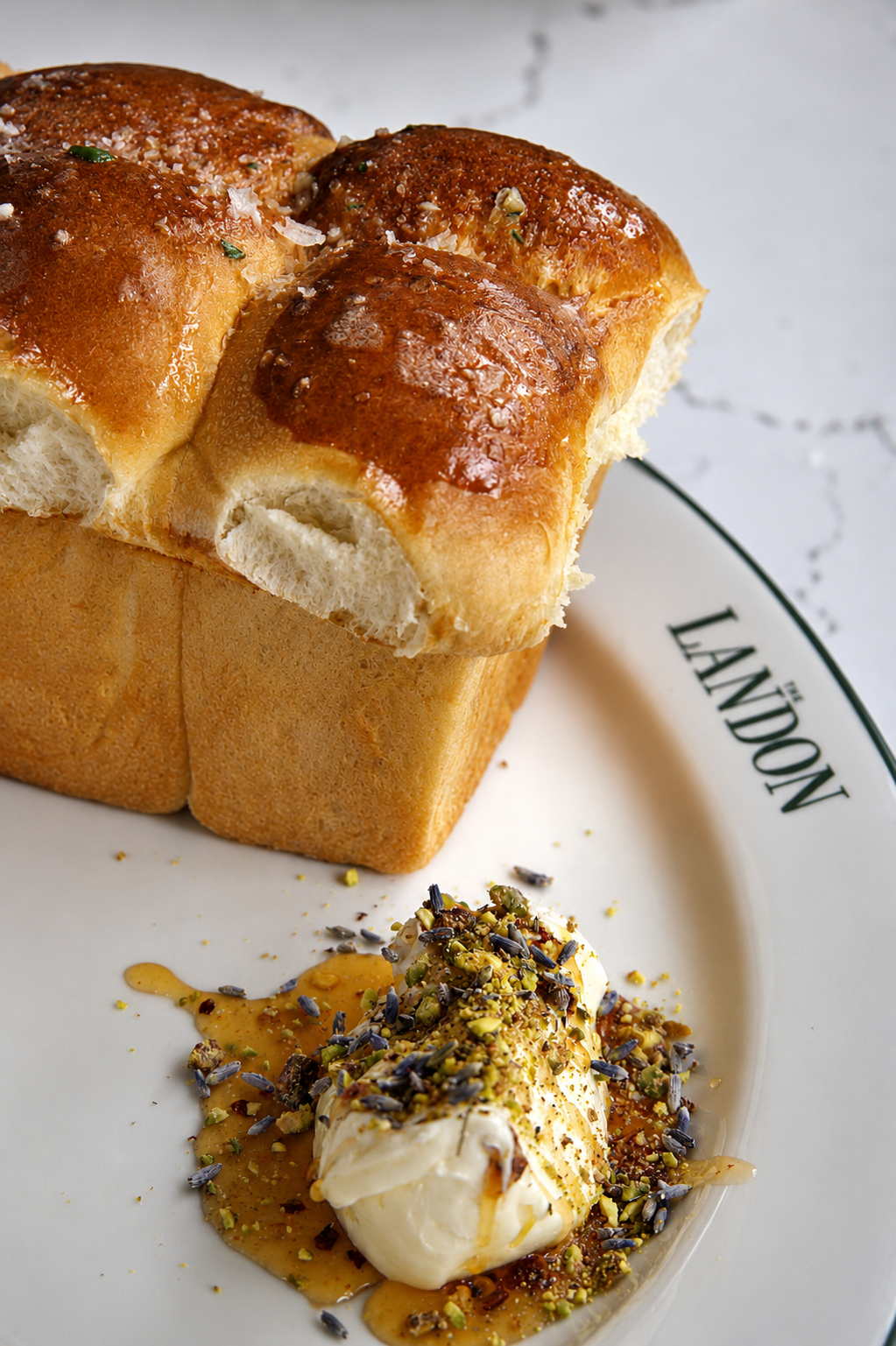 Three pieces of bread topped with melted cheese and herbs on a plate, served with a dollop of cream cheese, honey, chopped pistachios, lavender, and a drizzle of honey.
