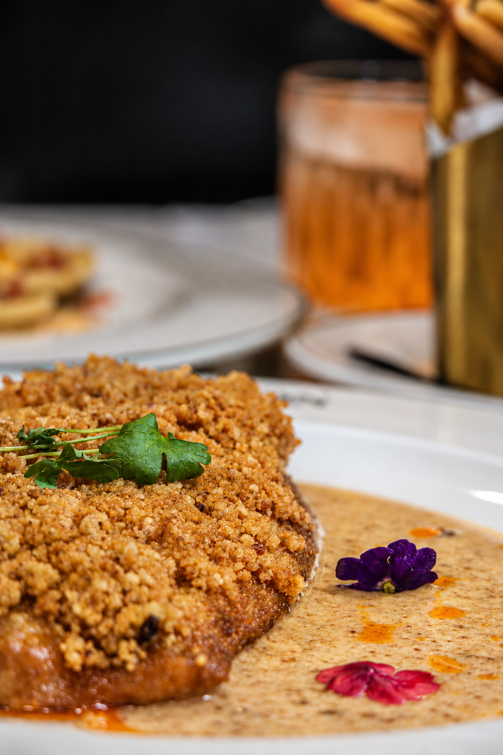 Breaded meat topped with herbs and served with a creamy sauce garnished with edible flowers on a white plate.