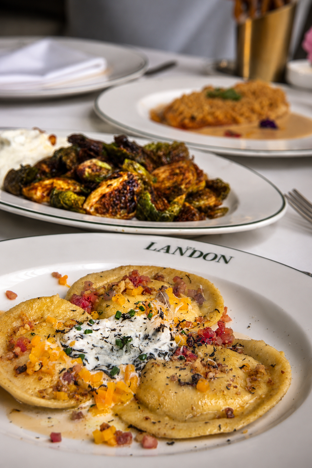 Plate of ravioli topped with cheese, bacon, and herbs in the foreground, with roasted chicken and vegetables in the background, on a dinner table.
