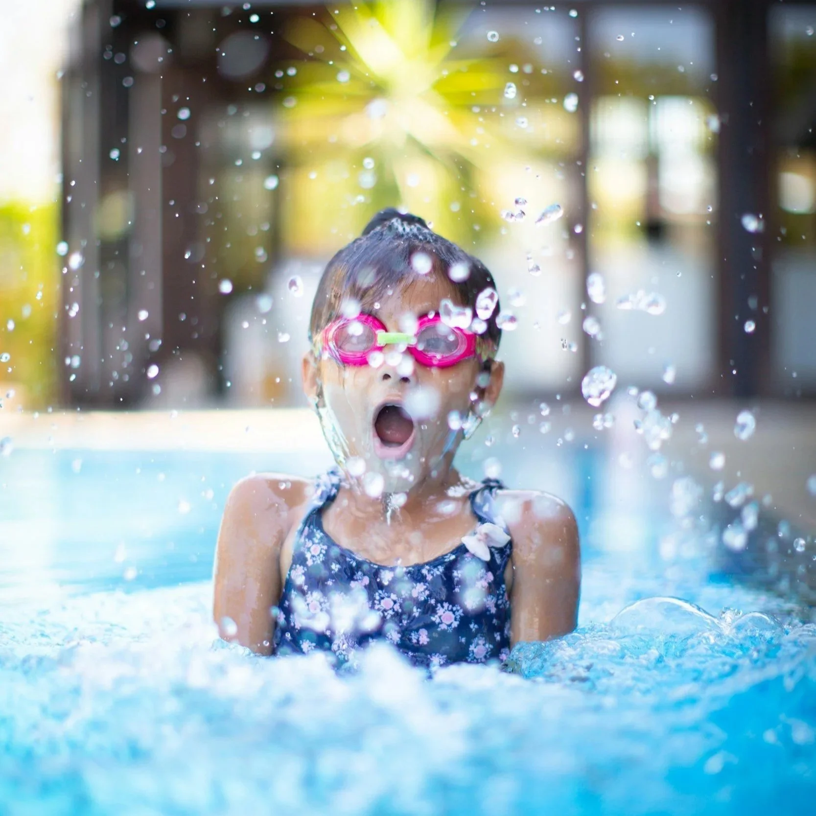 Child girl swimming in pool wearing pink goggles and a blue floral swimsuit, water splashing around her, background with bright natural light