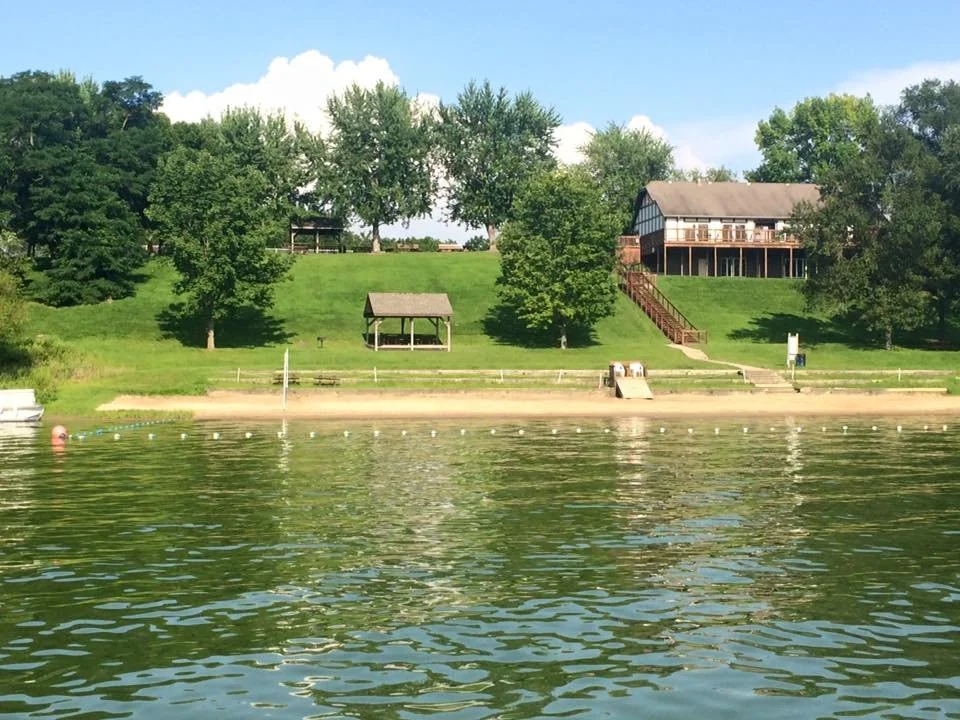 A lakeside with a grassy slope, trees, a small pavilion, stairs leading to a large house with a deck, and a sandy beach area.