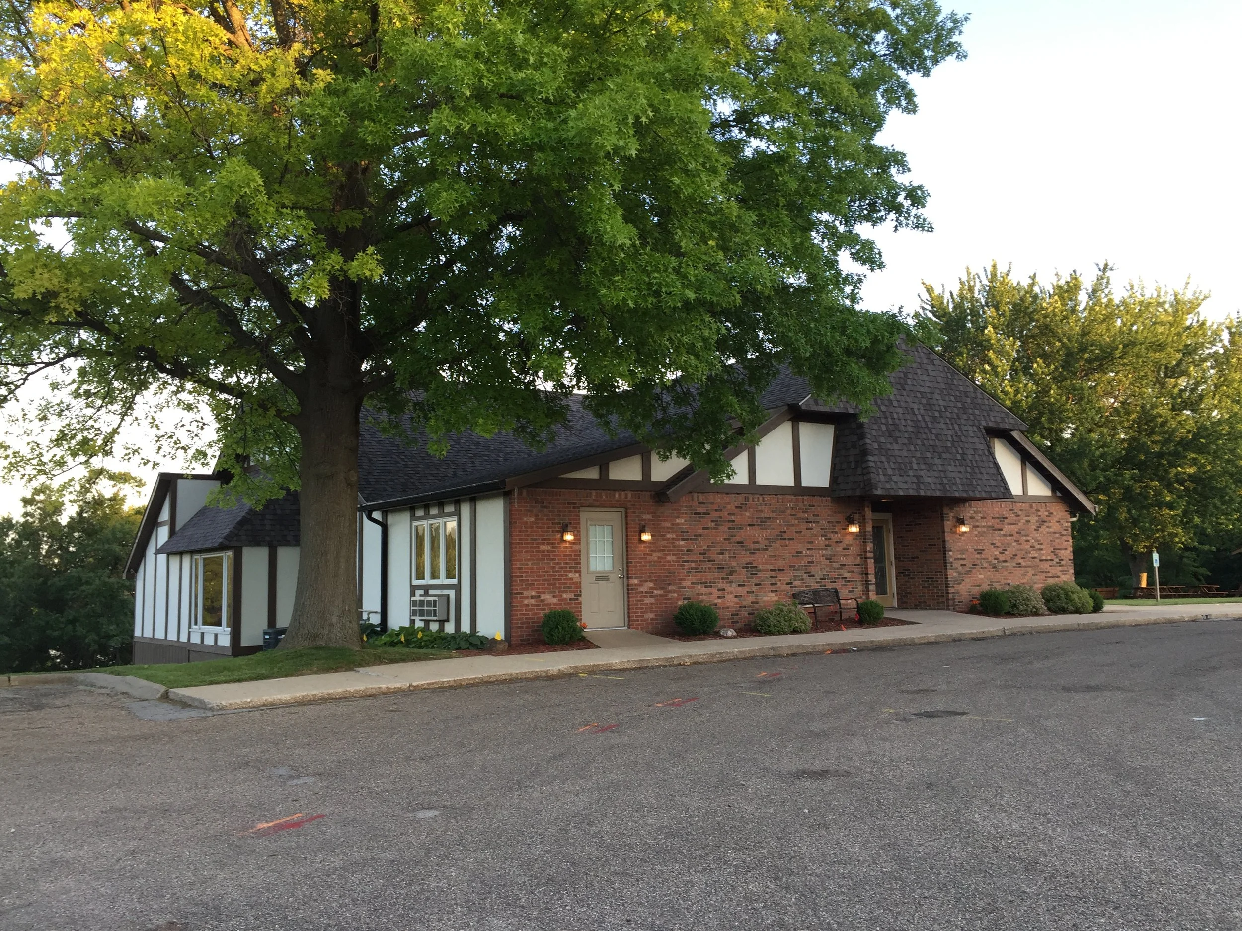 A residential building with brown brick walls and a dark shingle roof, situated under a large green tree. The house has several windows, a door with a small porch light, and a garden bed with shrubs around its perimeter. The scene is captured during early evening or late afternoon.