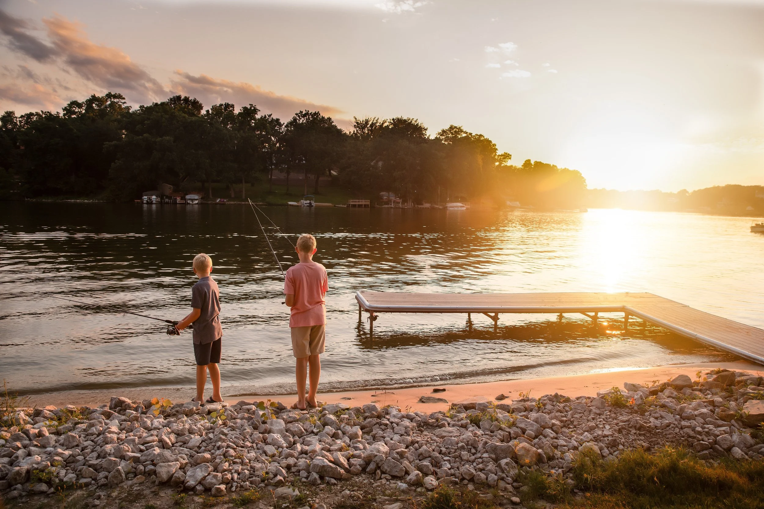 Two children fishing by a lake at sunset, with boats and trees along the shoreline and a rocky beach area in the foreground.