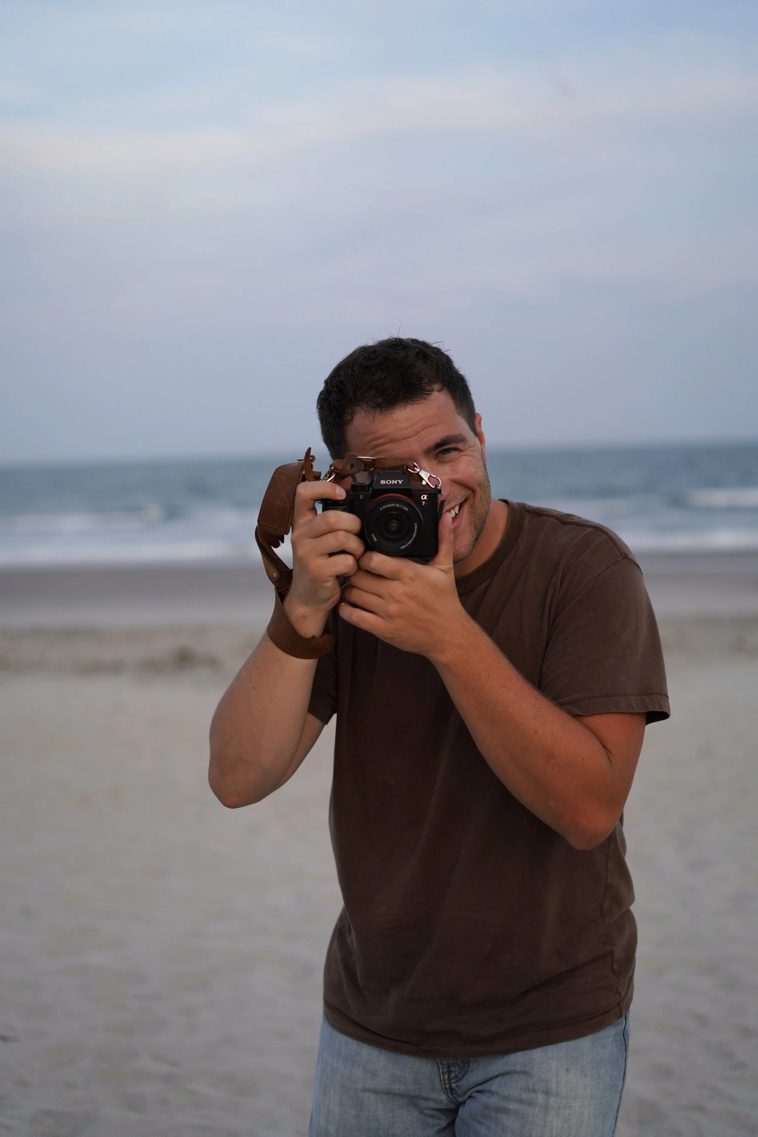 A smiling man with short dark hair taking a photo on a beach with the ocean and cloudy sky in the background.