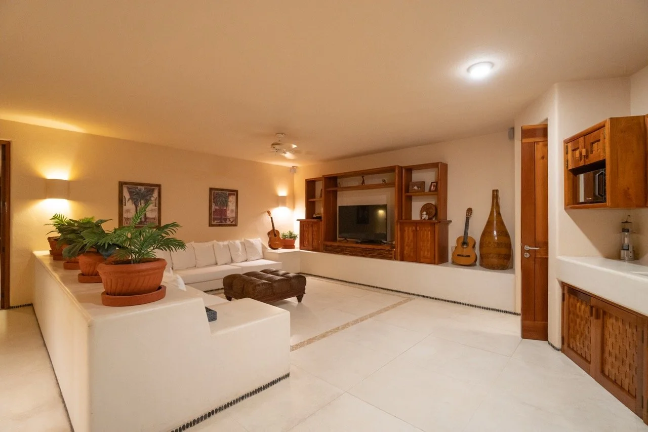Sala de estar con sillón blanco, plantas en macetas de barro, guitarras, una gran unidad de mueble de madera con televisión, y decoración en tonos cálidos.