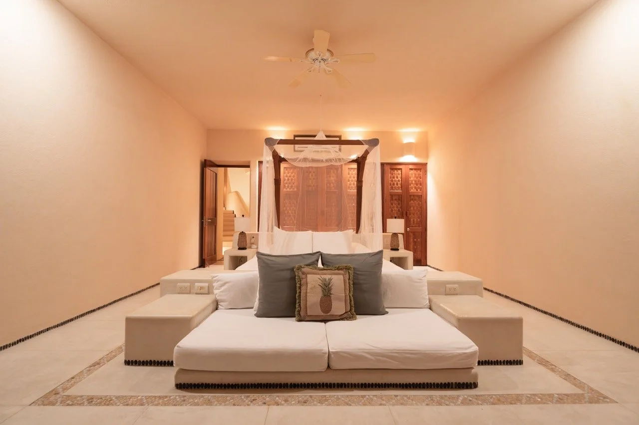 Habitación con cama grande y dos almohadas grises, una almohada decorativa con piña,, cortinas blancas, lámparas de noche, paredes de color claro y suelo con borde decorativo.