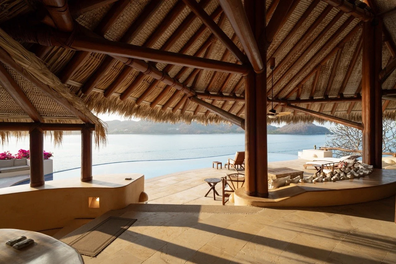 Vista desde una terraza con techo de paja, vista al mar, con sillas y mesas de madera, decoración de piedra y flores, en un lugar de descanso junto a la playa.