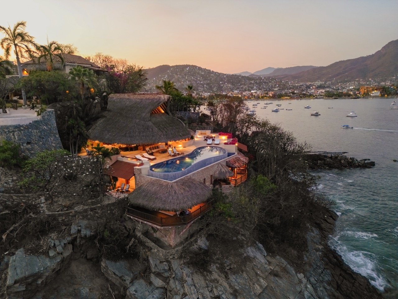 Vista aérea de una villa frente al mar con piscina y áreas de descanso, en un entorno de colinas y ciudad al atardecer.