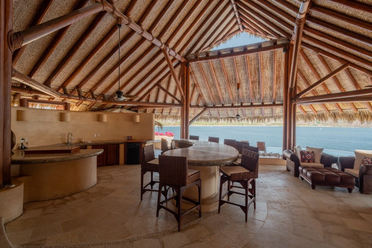 Área de descanso y cocina con vista al mar, techo de paja, mobiliario de madera y sillones, en una terraza o palapa junto a la playa.