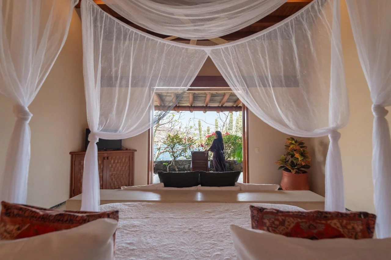 Vista del interior de una habitación con una cama con cabecera, cortinas blancas y una vista hacia un área exterior con plantas y una mujer de espaldas.