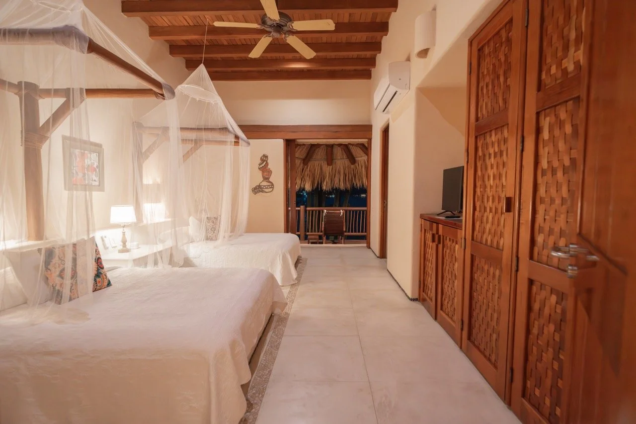 Habitación con dos camas con mosquiteros, una luz de noche, un ventilador en el techo, paredes blancas, armario de madera y una pequeña terraza con vista a una palapa y sillas.