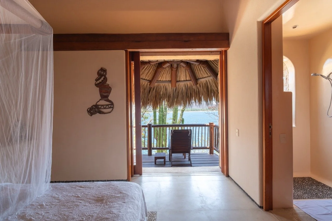 Habitación con vista a una terraza con silla reclinable y sombrilla de paja, vista a un lago o río, decoración rústica con madera y paredes claras.