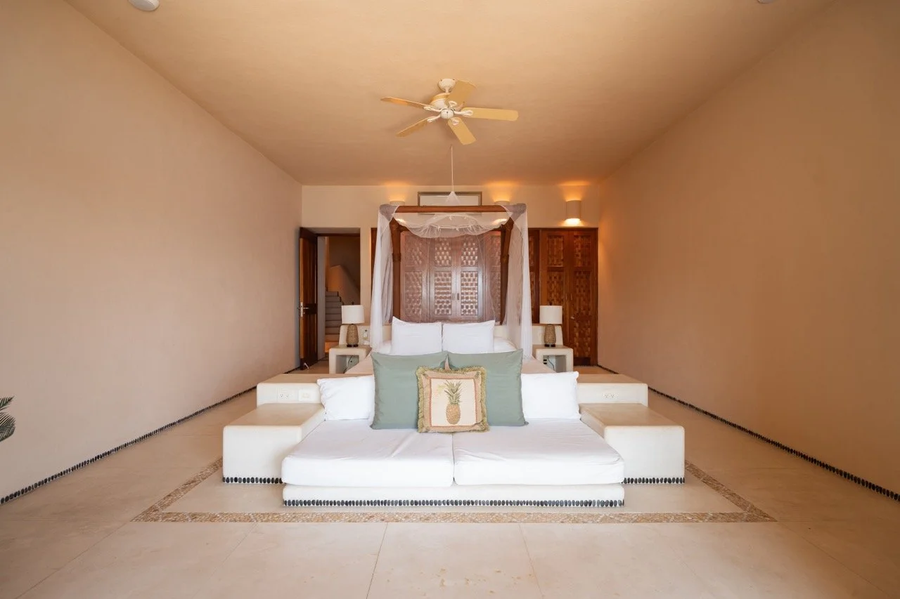 Habitación con cama grande y somier con dosel, sillones, alfombra y decoración en tonos claros con detalles de piña en cojines.