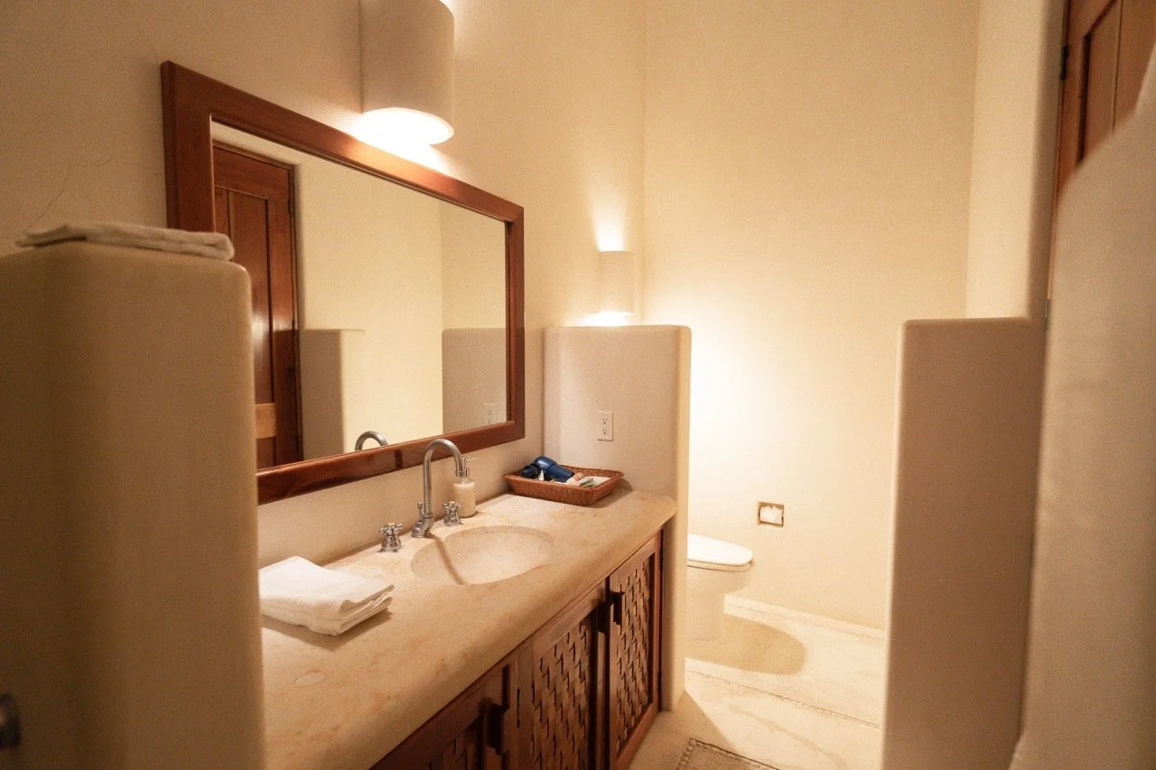 Baño con espejo, lavabo de piedra, toallas blancas y sanitarios en el fondo.