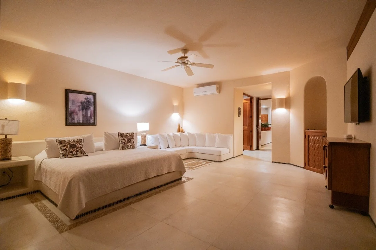 Sala de estar con cama grande, sofá blanco, lámparas y decoración sencilla, piso de baldosas claras, ventilador en el techo, aire acondicionado y televisión.