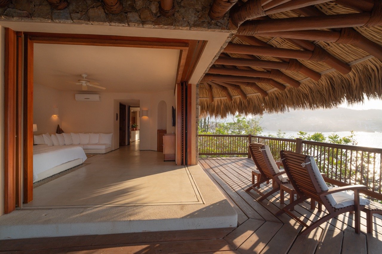 Amplia habitación de hotel con cama blanca y paredes blancas, vista desde la terraza de madera con techo de paja, con dos sillones de madera y vistas al río y la vegetación.
