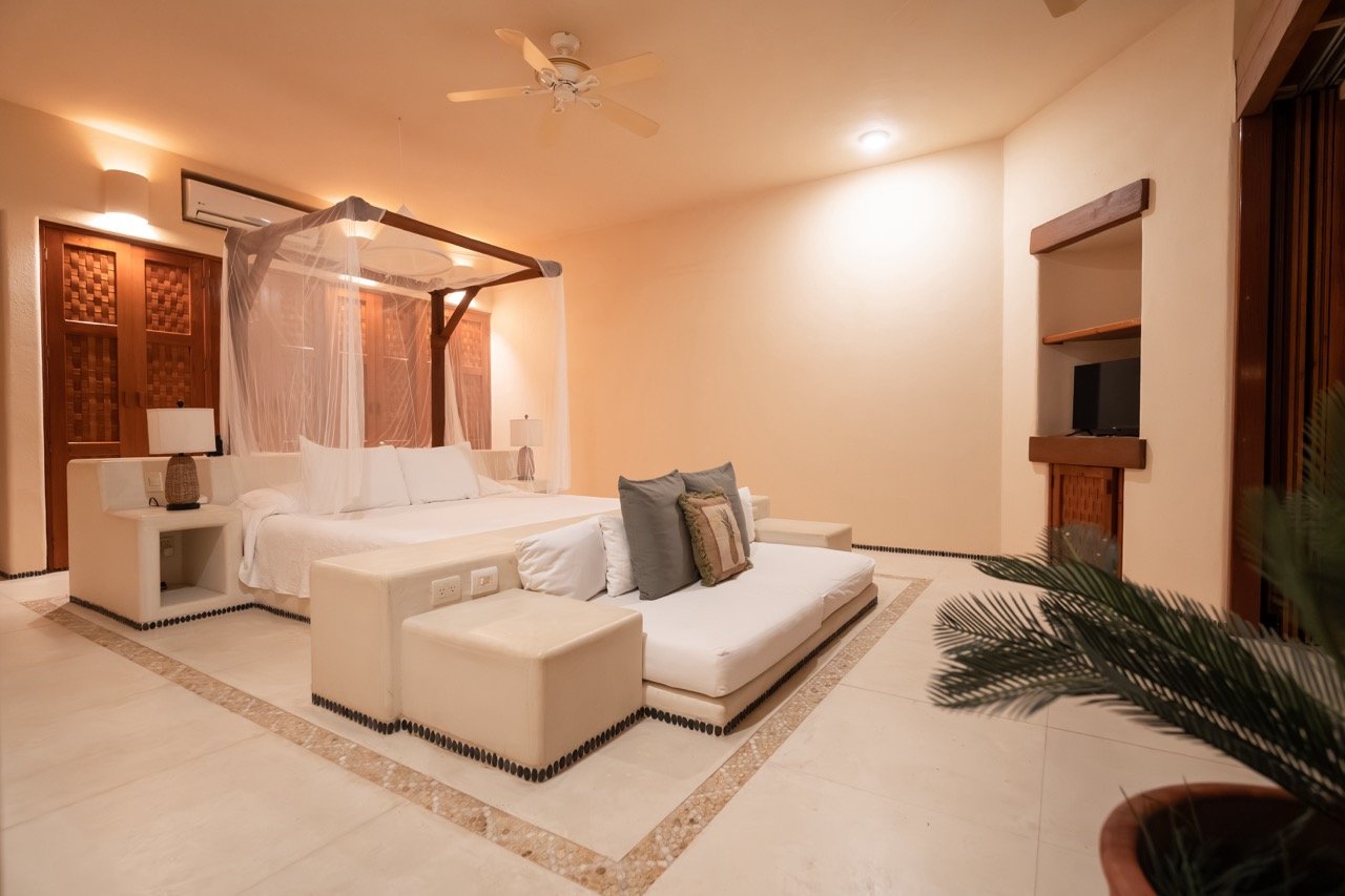 Habitación de hotel con cama grande con dos lámparas a cada lado, sillón blanco con cojines, paredes de color beige y decoración de madera.