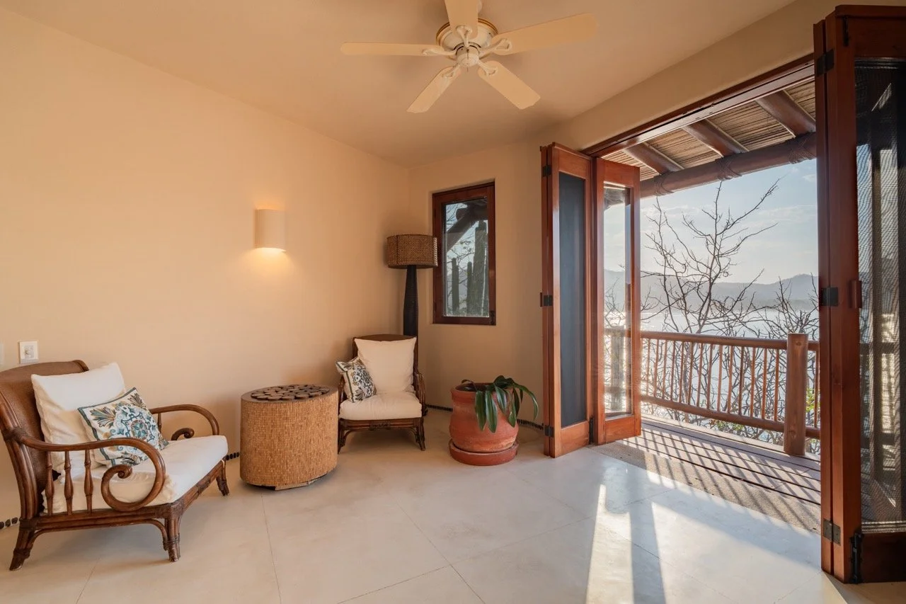 Sala con muebles de madera, ventana y puertas de madera abiertas al balcón con vista a árboles y montañas