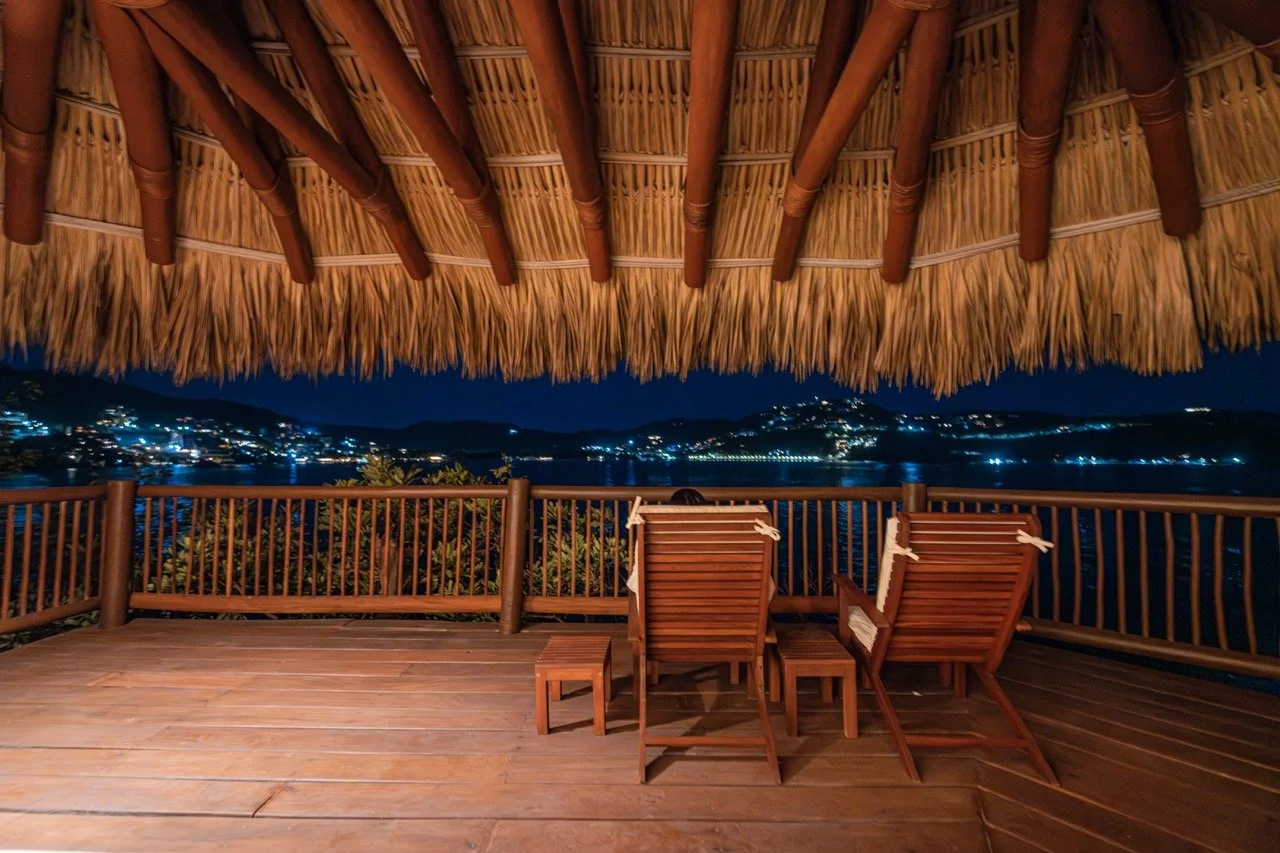 Vista nocturna desde una terraza de madera con sombrilla de paja y varias sillas, hacia un paisaje de montañas y ciudades iluminadas en la distancia.