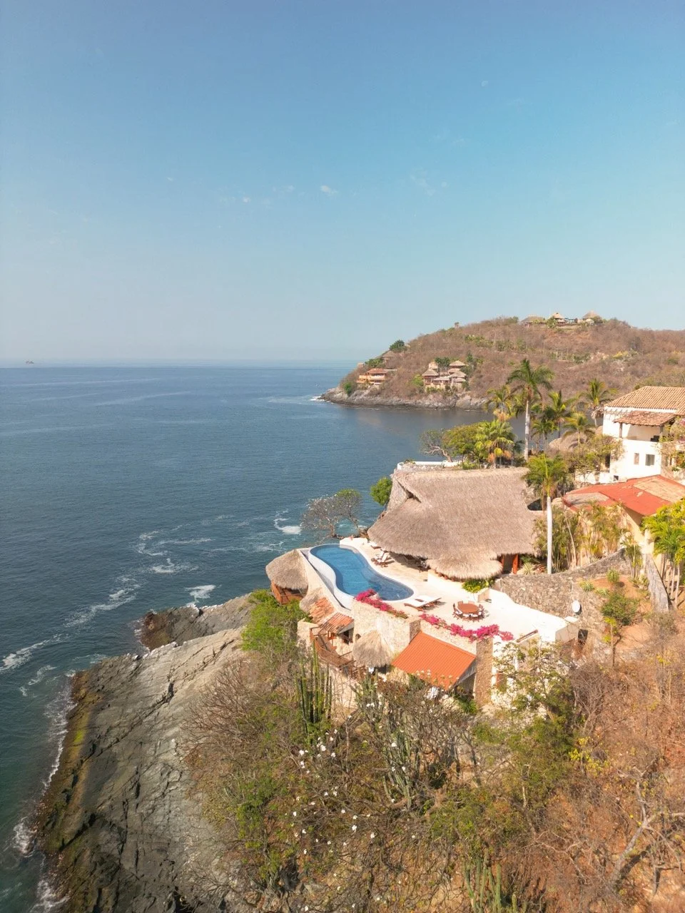 Vista aérea de una casa con piscina en la costa, rodeada de árboles y que mira al mar en un paisaje de colinas con más casas en el fondo.