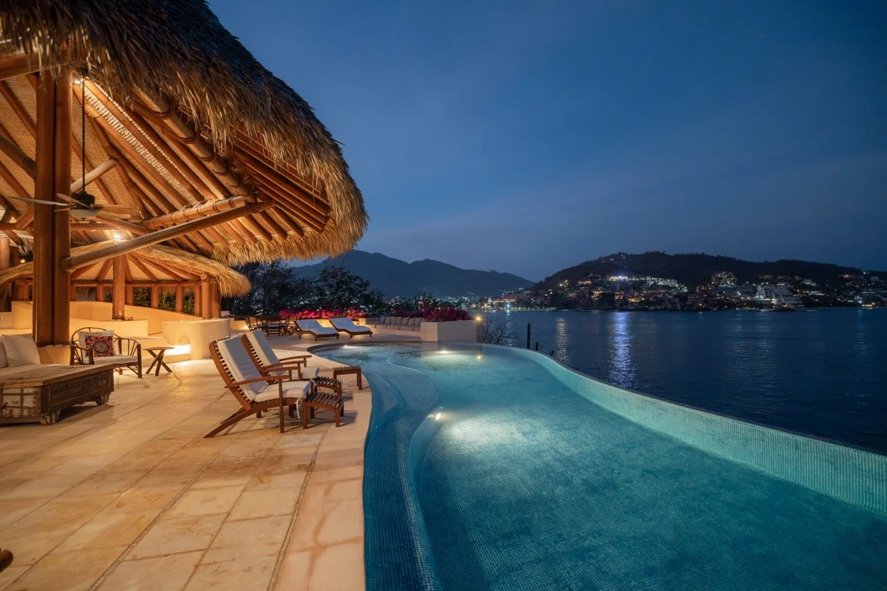 Vista nocturna de una piscina en un lugar de entrenamiento tropical, con cabañas de techo de paja y una vista del lago y las montañas al fondo.