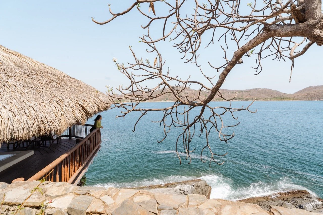 Una vista del mar con una palapa de techo de paja a la izquierda, una mujer mirando hacia el agua en la terraza, árboles sin hojas en primer plano y colinas en el fondo.