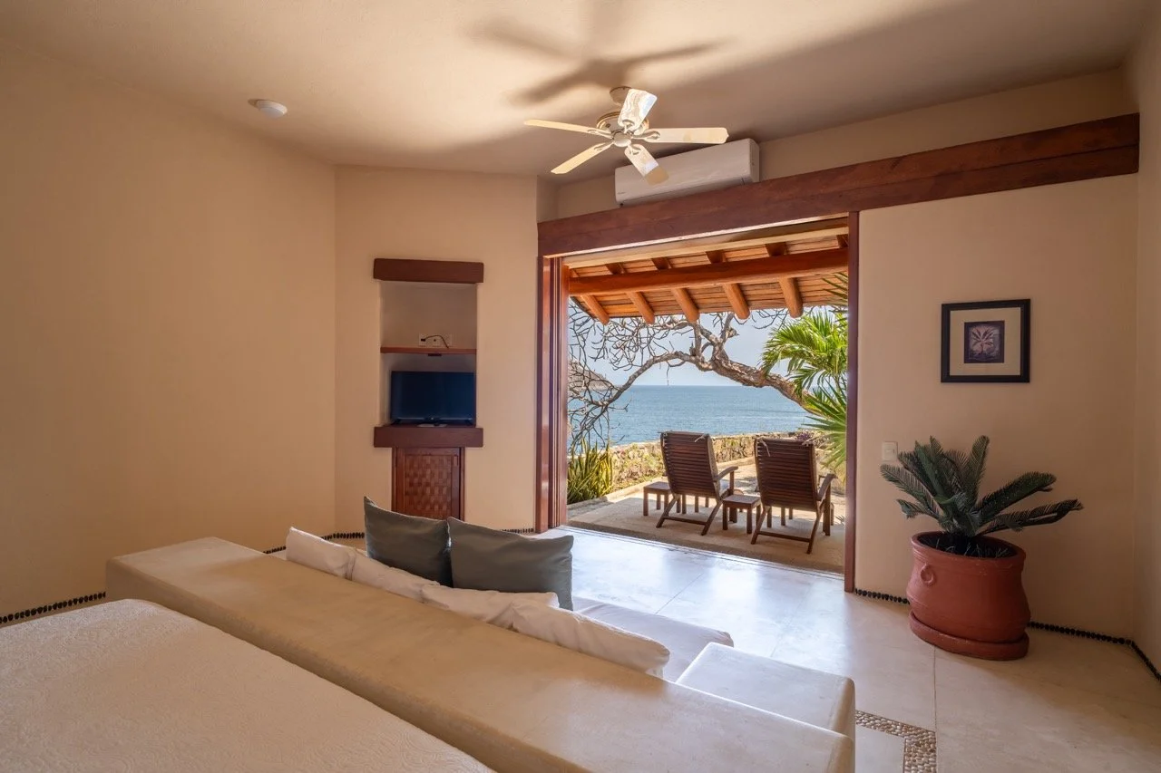 Habitación con vista al mar, balcón con silla y mesa, plantas y paredes beige.